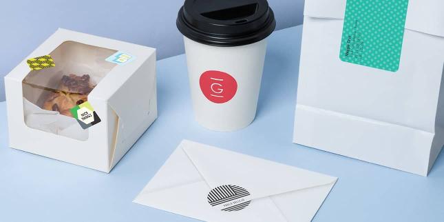 White pastry box, takeaway cup, envelope and paper pouch decorated with various colorful stickers on a light blue background