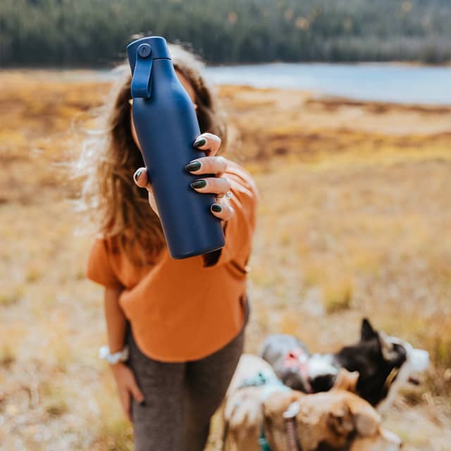 Woman with two dogs in a field holding a blue reusable water bottle by MOO